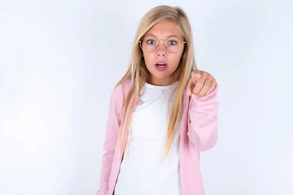 金发小女孩穿着粉色夹克 眼镜罩在白色的背景上 手指突兀地走在前面 张大了嘴 惊讶地看着前面的什么东西 — 图库照片