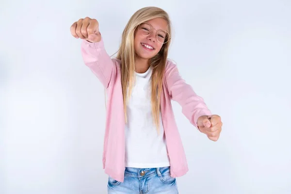 ホワイトバックグラウンド上のピンクのジャケットとメガネを身に着けているブロンドの少女が拳を示す — ストック写真