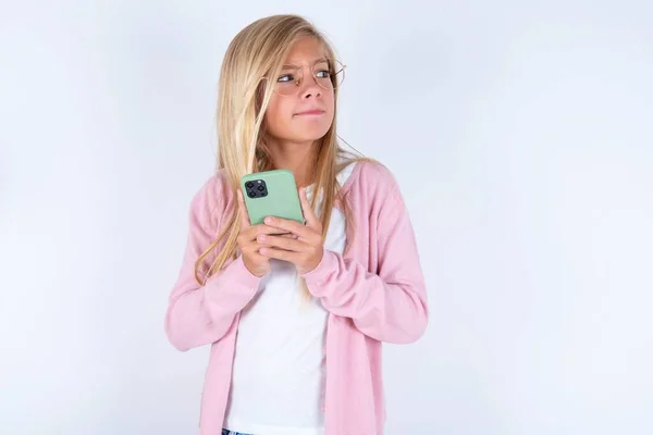 金发小女孩拿着智能手机 穿着粉色夹克 戴着一副白色背景的眼镜 — 图库照片