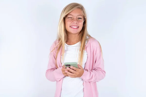 金发小女孩 身穿粉色夹克 戴着眼镜 头戴白色背景 手持智能手机 面带微笑 — 图库照片