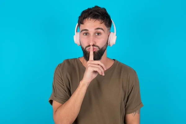 青い背景に立った若いヒスパニックのひげ付き男は 無線ヘッドフォンを着用して唇に指で隠しジェスチャーを作ります 静かに — ストック写真