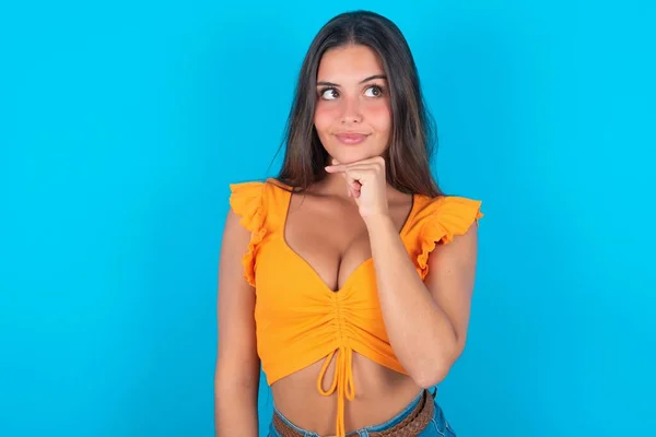Dreamy beautiful brunette woman wearing orange tank top over blue background with pleasant expression, looks sideways, keeps hand under chin, thinks about something pleasant.