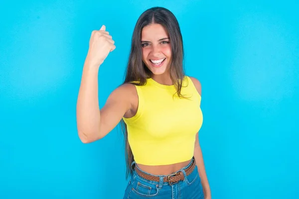 Fierce beautiful brunette woman wearing yellow tank top over blue background holding fist in front as if is ready for fight or challenge, screaming and having aggressive expression on face.
