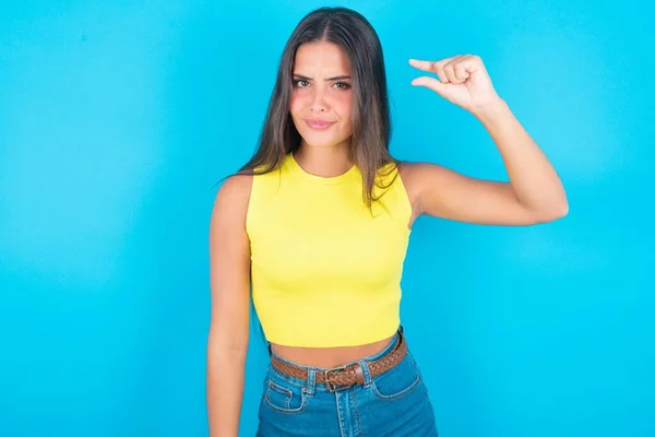 Upset beautiful brunette woman wearing yellow tank top over blue background shapes little gesture with hand demonstrates something very tiny small size. Not very much