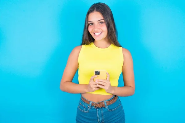 微笑着美丽的黑发女人 穿着黄色的背心 配上蓝色的背景 友善的 快乐的拿着手机 在镜子里自拍 — 图库照片