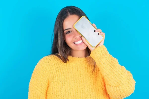 年轻的黑发女子 身穿蓝色背景的黄色毛衣 手持现代智能手机 一边微笑一边捂住一只眼睛 — 图库照片