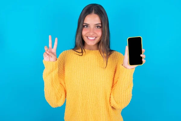 年轻的黑发女子 身穿蓝色背景的黄色毛衣 手持现代智能手机 并展示V字形标志 — 图库照片