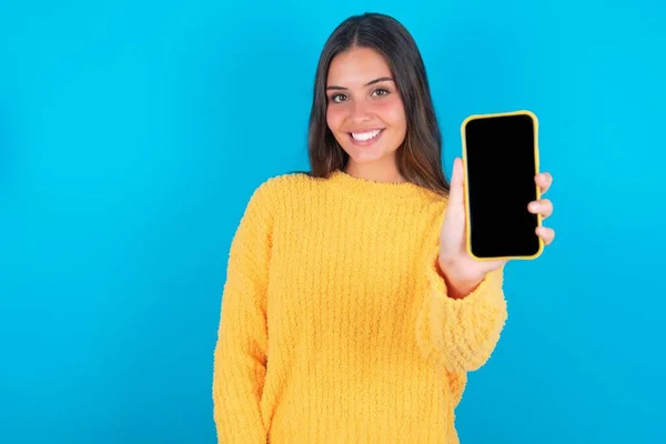 迷人的年轻黑发女子 穿着黄色毛衣 蓝色背景 手持现代设备 显示黑色智能手机黑色屏幕 — 图库照片