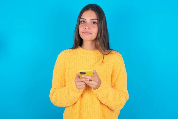 严肃而自信的年轻黑发女人手持电话 穿着黄色毛衣 蓝色背景的画像 — 图库照片