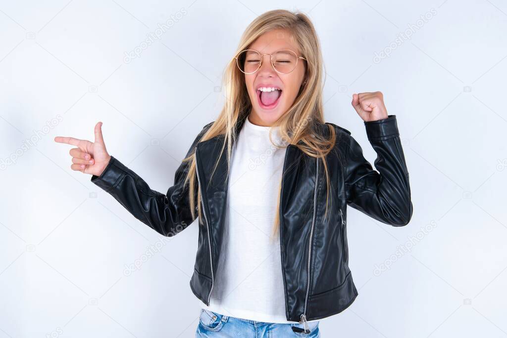 Cheerful beautiful blonde little girl wearing biker jacket and glasses over white background showing copy space and celebrating luck