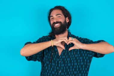 Mavi arka planda mavi tişörtlü, sakallı beyaz bir adam. Aşkla gülümsüyor. Elleriyle kalp sembolü ve şekli gösteriyor. Romantik konsept.