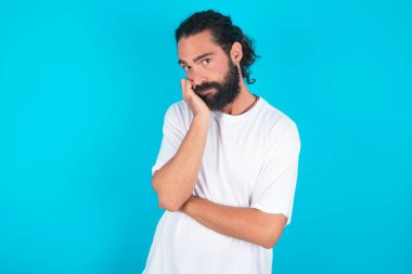 Çok sıkılmış, sakallı, beyaz tişörtlü, mavi arka planda beyaz tişörtlü beyaz bir adam, el ele tutuşurken, diğer eli çaprazlanmış, yorgun ve hasta görünüyor..