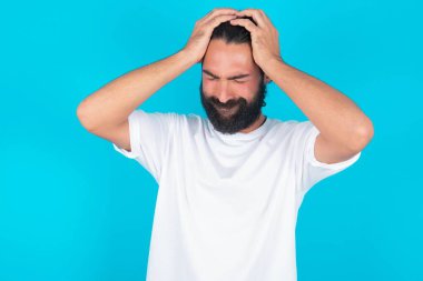 Beyaz tişörtlü beyaz sakallı beyaz bir adam mavi arka planda el ele tutuşmuş, baş ağrısı çekiyor, şakaklarına parmak bastırıyor.