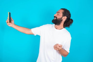 Beyaz sakallı beyaz tişörtlü beyaz bir adamın portresi mavi arka planda. Arkadaşlarına ve takipçilerine göndermek için selfie çekiyor ya da sosyal medyasına yolluyor..