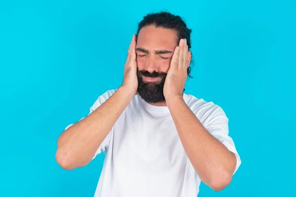 Caucasian man with beard wearing white T-shirt over white background holding head in hands with unhappy expression watching sad movie about animals and trying not to cry.