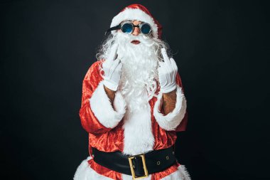 Man dressed as Santa Claus making a rude gesture, raise his fingers as a sign of 'fuck you', on black background. Christmas concept, Santa Claus, gifts, celebration.