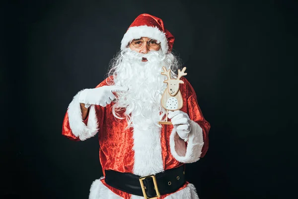 一个穿得像圣诞老人的人 用手指指着一只黑色背景的木制驯鹿 圣诞老人 庆典的概念 — 图库照片