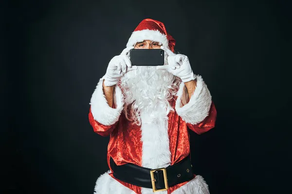 男人穿得像圣诞老人 手里拿着一部手机 拍了一张照片 背景是黑色的 圣诞老人 庆典的概念 — 图库照片