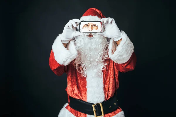 男人穿得像圣诞老人 手里拿着一部手机 拿着一个黑色背景的自拍 圣诞观念 圣诞老人 庆祝活动 — 图库照片