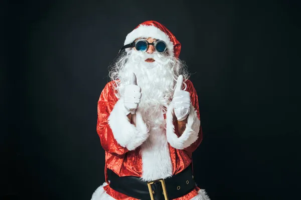 男人打扮成圣诞老人 戴着维多利亚风格的焊缝护目镜 竖起大拇指 背景是黑色的 圣诞老人 庆典的概念 — 图库照片