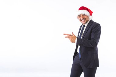 Takım elbiseli, kravat takmış, Noel Baba şapkası takmış gülümseyen, beyaz arka planda elleriyle işaret eden bir iş adamı. Noel kutlaması kavramı.