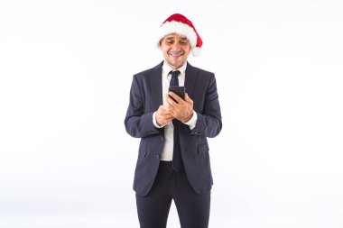 Takım elbiseli, kravatlı ve Noel Baba şapkalı bir işadamı beyaz arka planda cep telefonuna bakıyor. Noel kutlaması kavramı.