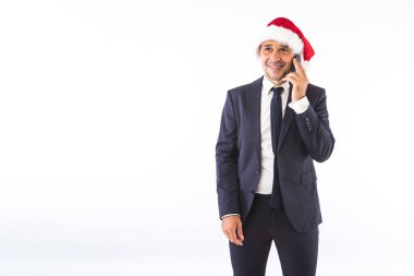 Takım elbiseli, kravatlı, Noel Baba şapkalı beyaz arka planda telefonda konuşan bir iş adamı. Noel kutlaması kavramı.