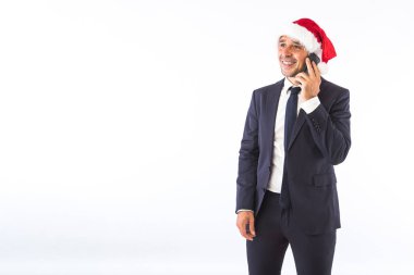 Takım elbiseli, kravatlı, Noel Baba şapkalı beyaz arka planda telefonda konuşan bir iş adamı. Noel kutlaması kavramı.