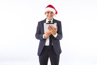 Takım elbiseli, kravatlı, Noel Baba şapkalı beyaz arka planda elinde tabletle gülümseyen bir iş adamı. Noel kutlaması kavramı.