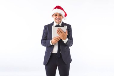 Takım elbiseli, kravatlı ve Noel Baba şapkalı bir iş adamı beyaz arka planda tabletine bakıp gülümsüyor. Noel kutlaması kavramı.