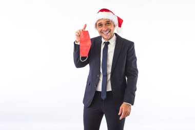 Takım elbiseli, kravatlı, Noel Baba şapkalı, beyaz arka planda kırmızı bir maske takan bir iş adamı. Noel kutlaması kavramı.
