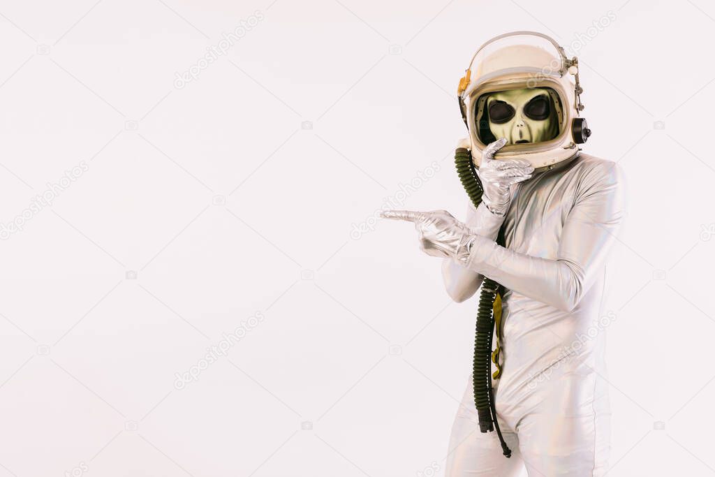Person dressed in silver suit and green alien mask, wearing a cosmonaut helmet and pointing with hand and finger, on white background