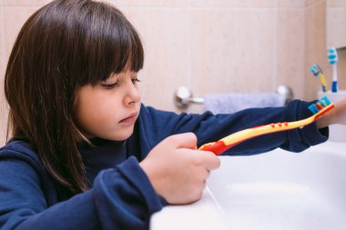 Koyu mavi kazaklı küçük bir kız, diş fırçasını lavaboda, banyoda tutuyor. Diş fırçalama, hijyen ve çocukluk konsepti.