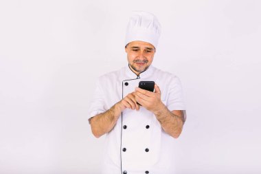 Beyaz şapkalı ve ceketli aşçı beyaz arka plandaki cep telefonuna bakıyor. Restoran, yemek ve paket servis konsepti..