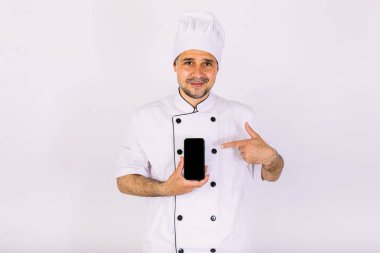 Beyaz şapkalı ve ceketli şef cep telefonunu beyaz arka planda gösteriyor. Restoran, yemek ve paket servis konsepti..