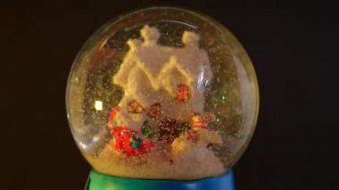 Noel Baba 'nın olduğu cam bir kar küresi, bir ren geyiği ve içinde yansıyan renkli ışıklarla hareket eden karlı bir ev. Noel, Noel Baba ve kutlama konsepti.