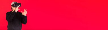 Kısa sakallı, siyah kazaklı, şapkalı ve sanal gerçeklik gözlüklü genç adam afişi kırmızı arka planda adeta bir şeye dokunuyor. Teknoloji, VR, hesaplama ve hobiler.