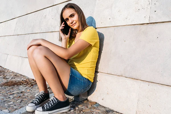 Sarı tişörtlü genç bir kız akıllı telefonuyla taş duvara yaslanmış bir şekilde konuşuyor. Teknoloji, müzik, yaşam tarzı ve rahatlama kavramı.