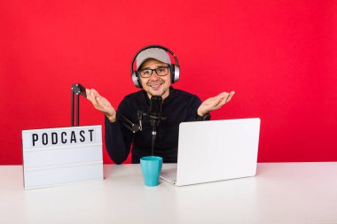 Podcast radyo kayıt stüdyosunda kapaklı bir sunucu el hareketleri yapıyor, bir bilgisayarın yanında, bir mikrofon ve podcast 'li bir ışık kutusu, kırmızı arka planda. Podcast, yayın konsepti