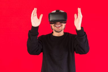 Siyah giyinmiş, sanal gerçeklik gözlüğü takan bir adam kırmızı arka plandaki bir şeye dokunurken gülümsüyor. Teknoloji, VR, hesaplama ve hobiler.