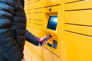 Sarı dolaptan bir paket almak için cep telefonundaki kodu tarayan bir adamın kolu. Mesajlaşma kavramı, çevrimiçi, e-ticaret ve paketleri karşılaştırın