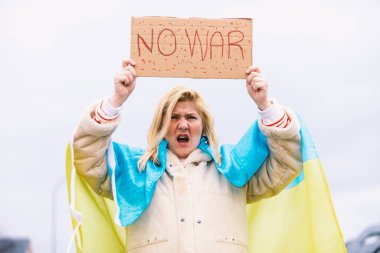 Ukraynalı sarışın ve ciddi jestli bir kadın, mavi ve sarı bir Ukrayna bayrağıyla sokakta Ukrayna-Rusya Savaşı 'nı protesto eden bağırıyor..