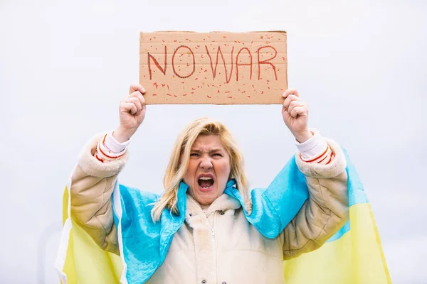 Ukrainian woman with blonde hair and serious gesture, with a blue and yellow Ukrainian flag with a sign that says: 'No War', in the street, shouting protesting the Ukraine-Russia War.