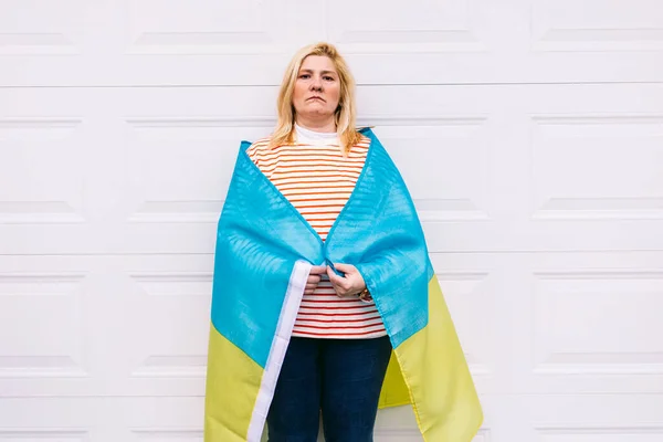 Ukrainian mature woman with blond hair serious gesture with blue and yellow Ukrainian flag on white background in the street. Ukraine war, invasion, protest, patriotism and no war concept.
