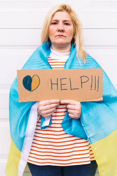 Ukrainian woman with blonde hair and a serious gesture, with a blue and yellow Ukrainian flag holding a sign with a yellow and blue heart that reads: \'Help\', on the street. Concept Ukraine war, invasion, protest, patriotism.