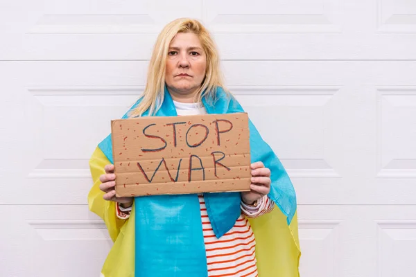 Ukrainian woman with blonde hair and a serious gesture, with a blue and yellow Ukrainian flag with a sign that reads: \'Stop War\', on the street. Concept Ukraine war, invasion, protest, patriotism.