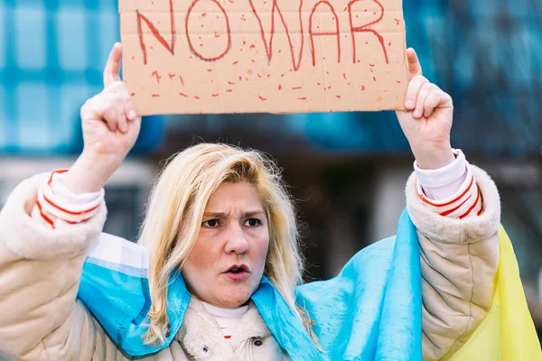 Ukrainian woman with blonde hair and serious gesture, with a blue and yellow Ukrainian flag with a sign that says: \'No War\', in the street, shouting protesting the Ukraine-Russia War.