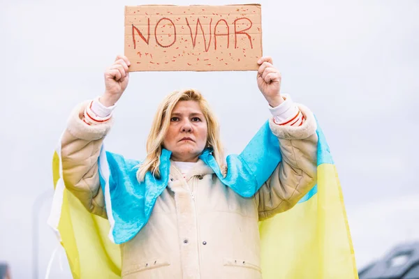 Ukrainian woman with blonde hair and serious gesture, with a blue and yellow Ukrainian flag with a sign that says: \'No War\', in the street, protesting the Ukraine-Russia War.