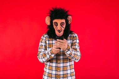 Şempanze maskeli ve ekose gömlekli bir adam kırmızı arka planda cep telefonuna danışıyor..