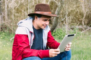 Girişimci serbest çalışan şapkalı bir adam, sahada dinlenirken tabletine bakıyor, bir taşın üzerinde oturuyor. İş kavramı, keyif, rahatlama, İnternet, teknoloji ve bağlantı.